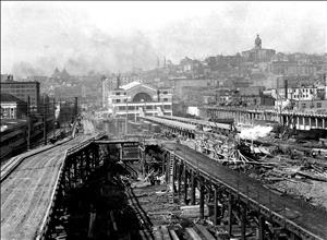 Roads on wooden trestles pass over a construction site where large timbers are stacked. A large brick building stands at the end of the construction area. City buildings cover the hillside in the background with several clocktowers and turrets visible on top of the hillside. 